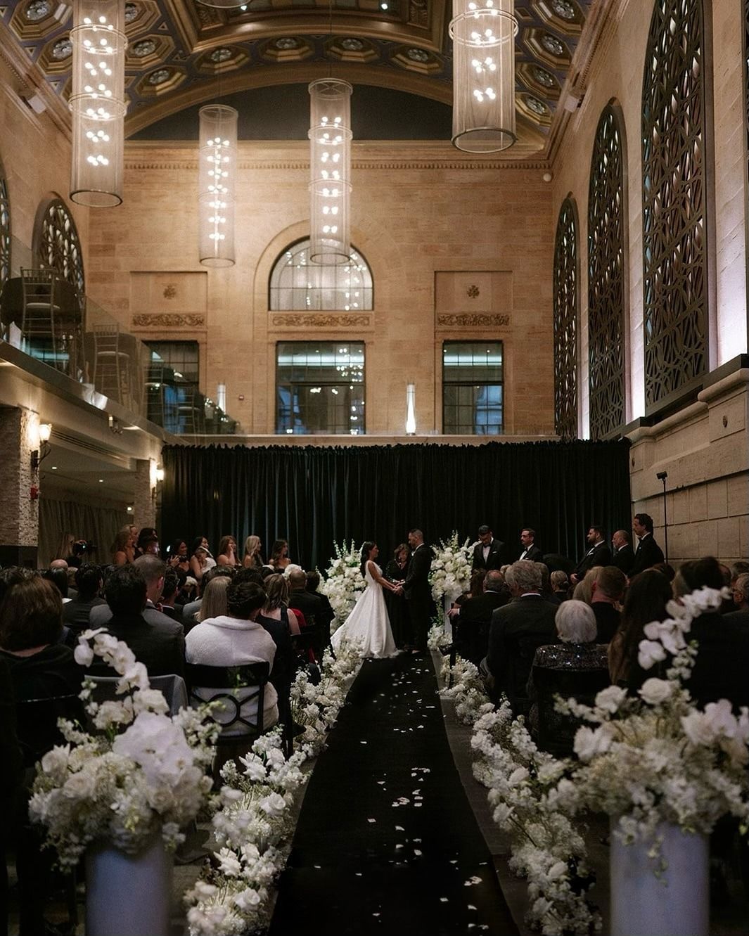 Wedding ceremony in a grand hall with white floral arrangements and a black aisle leading to the altar
