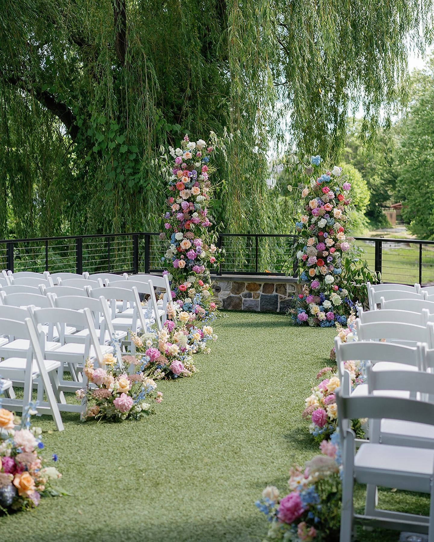 Outdoor wedding aisle with white chairs and pastel floral arches under a leafy tree.