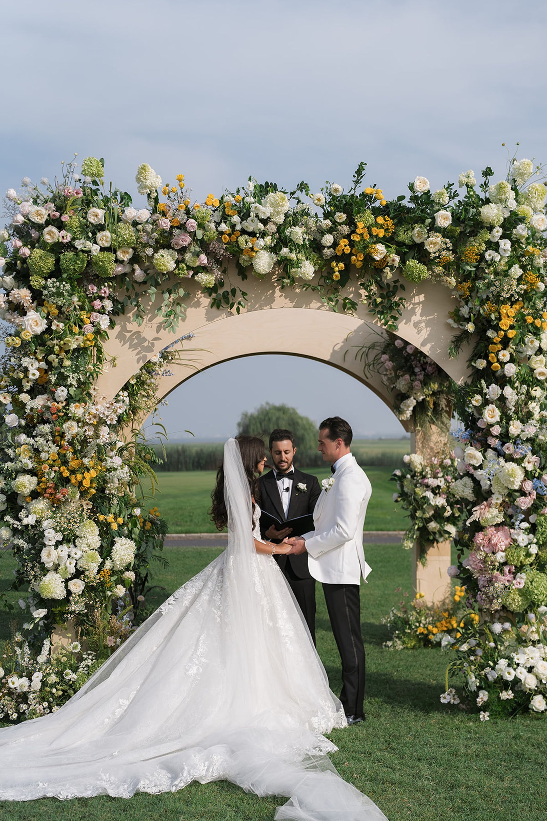 Wedding couple under a floral arch on a grassy lawn, holding hands, with a sea view in the background.