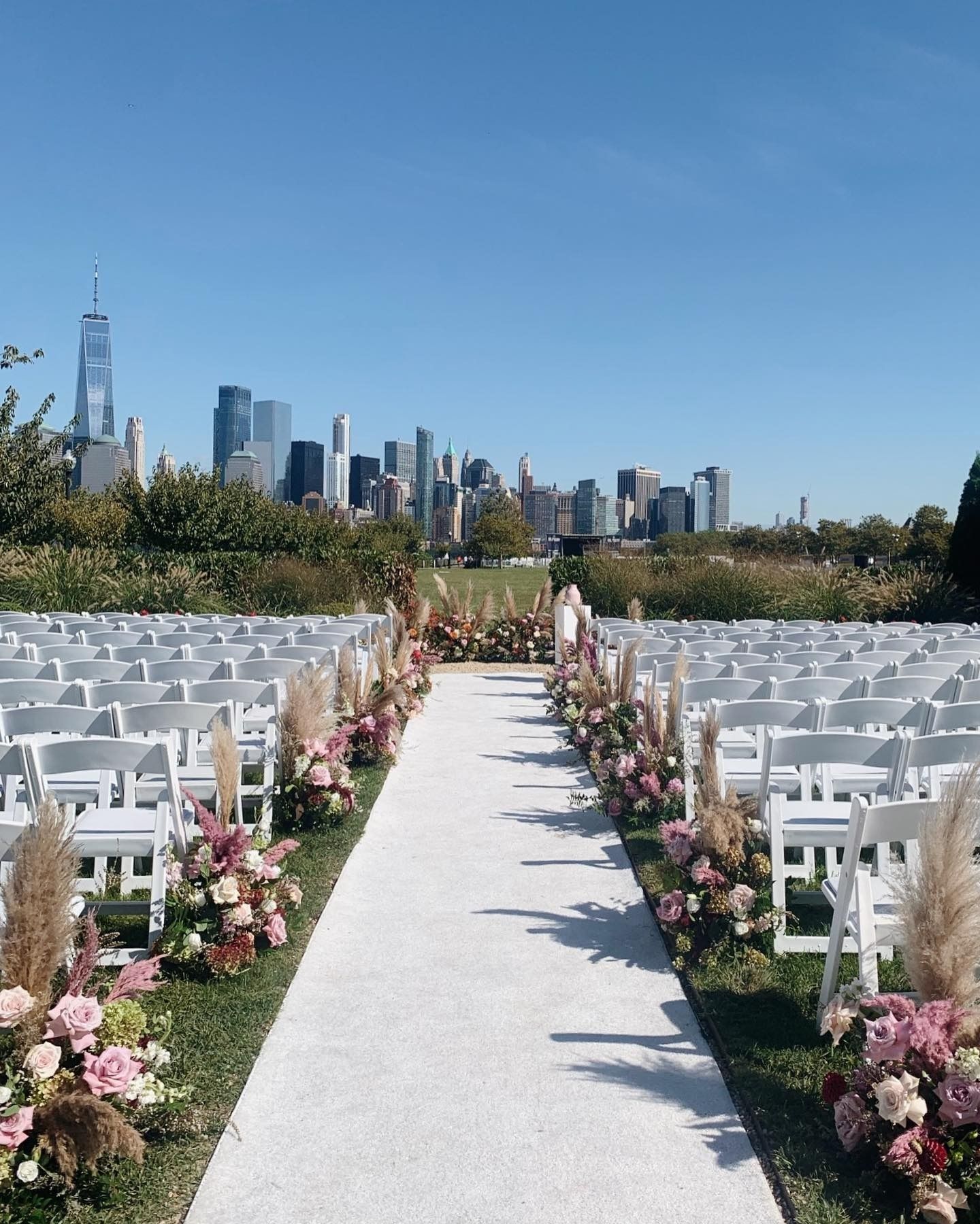 Outdoor wedding aisle lined with white chairs and pink flowers, with a city skyline in the background