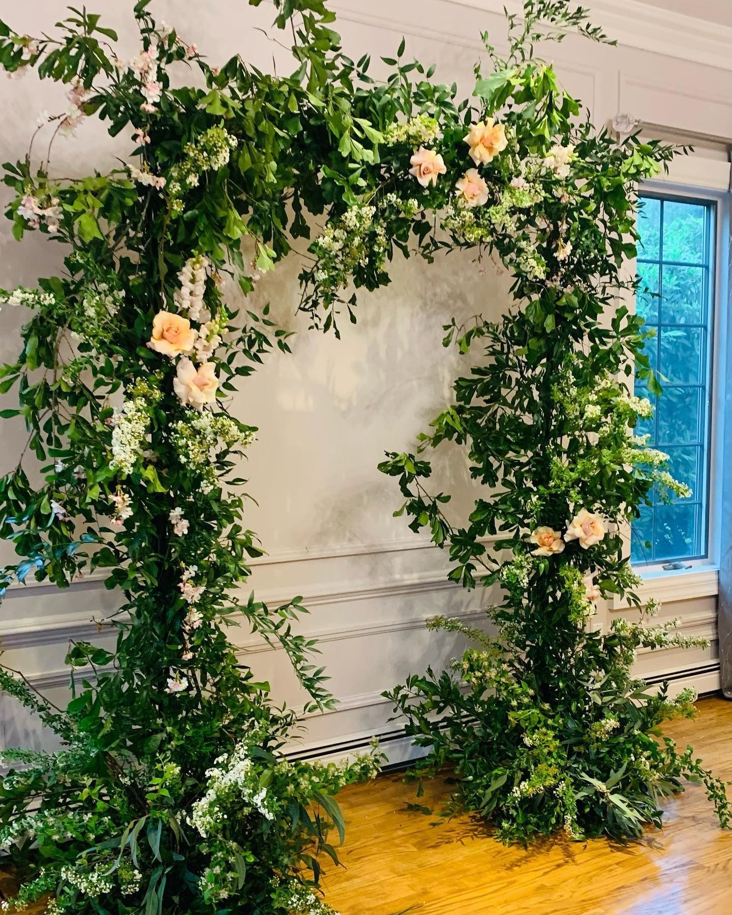 Green floral arch with pale pink flowers against a white wall in a bright indoor room