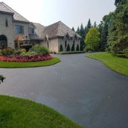 Large suburban house with a curved driveway, manicured lawn, and flower bed lined with evergreen trees