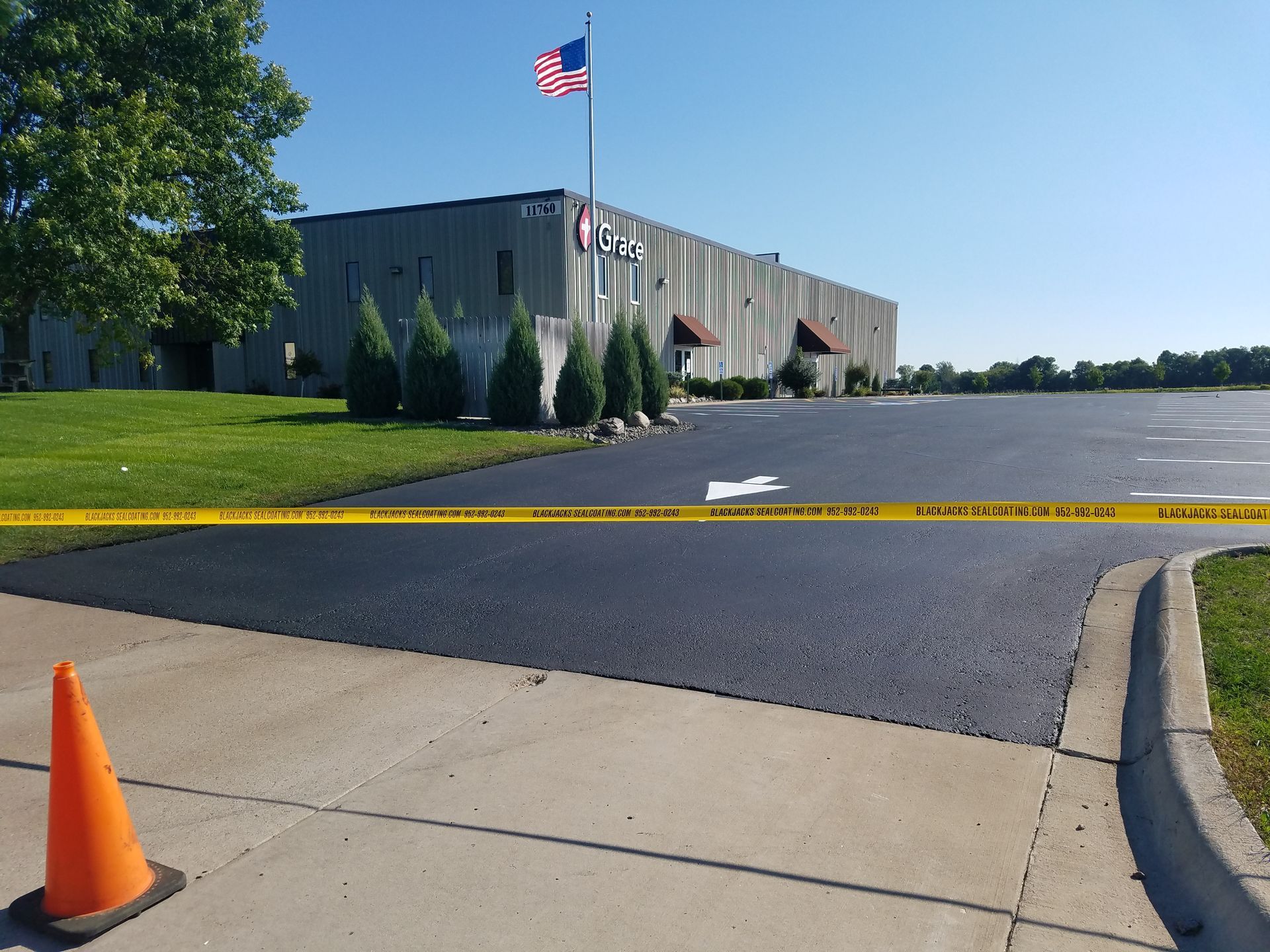 Yellow caution tape blocks a parking lot beside a gray building with an American flag and orange cone.