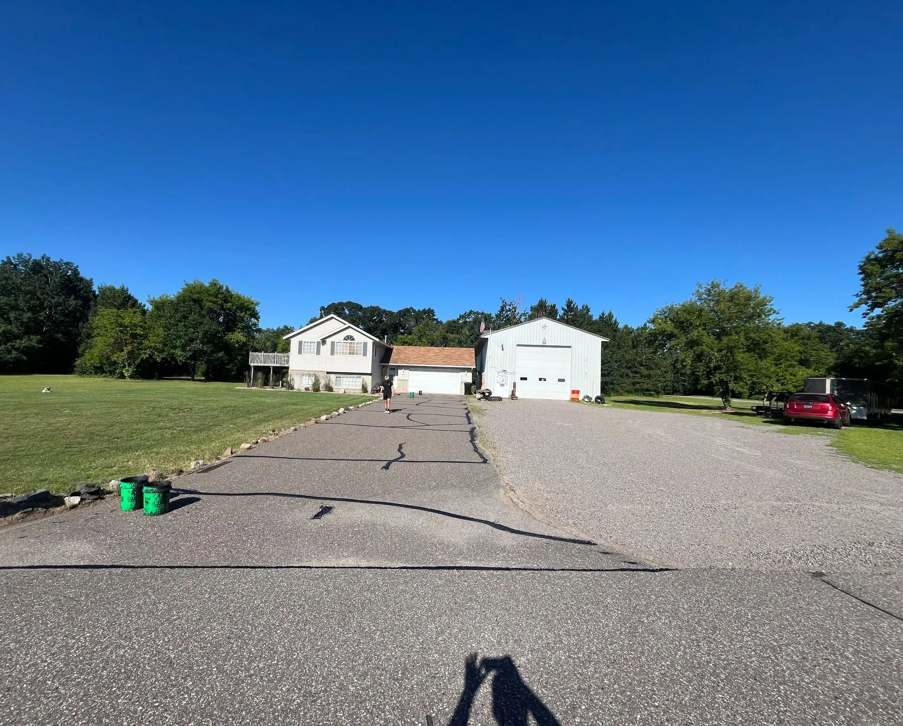 Gravel driveway leading to a house and garage under a clear blue sky, with trees and a red car on the right.