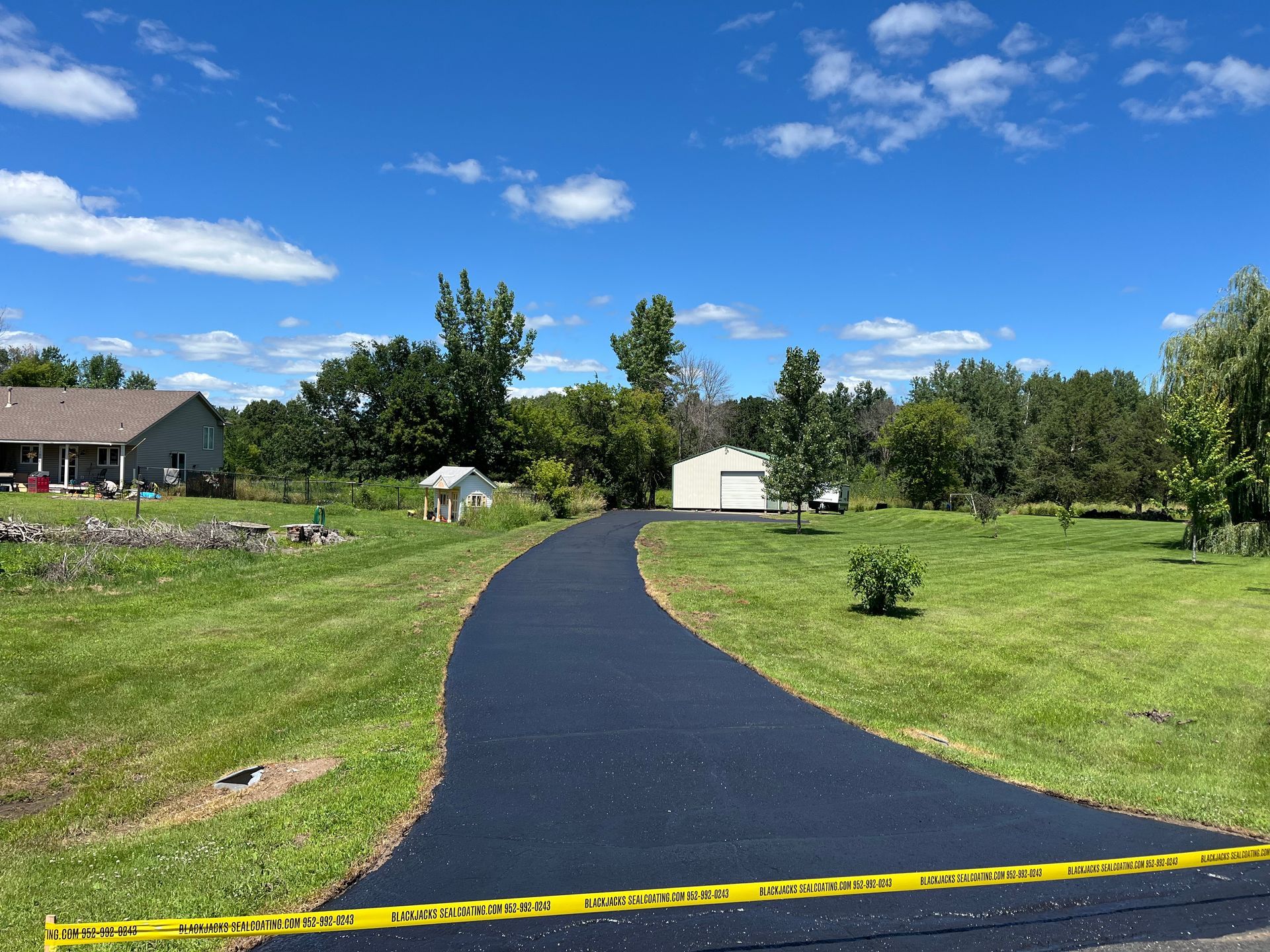 Curved black asphalt path through a green park under a blue sky, with a house and trees in the background