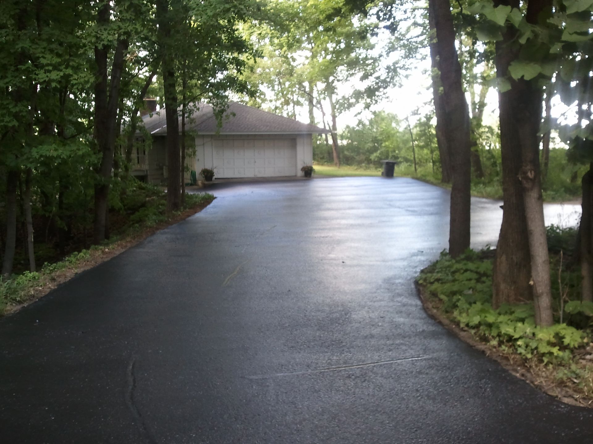 Wet paved driveway curving through trees toward a garage at the end of a wooded lot