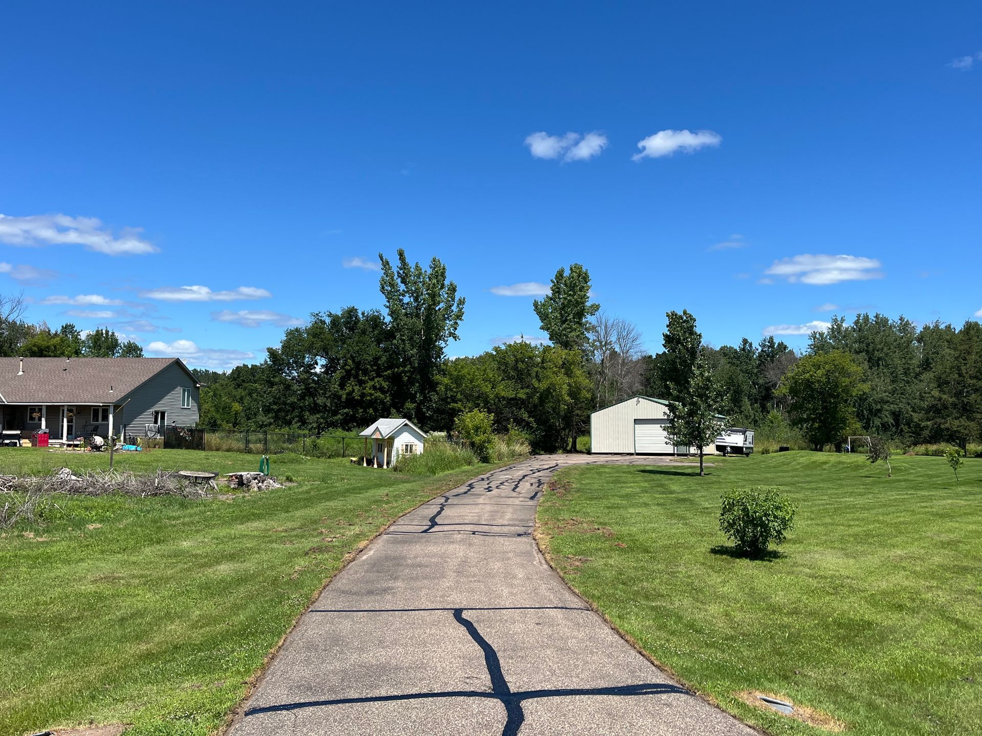 Cracked concrete path through a grassy yard toward small buildings under a blue sky