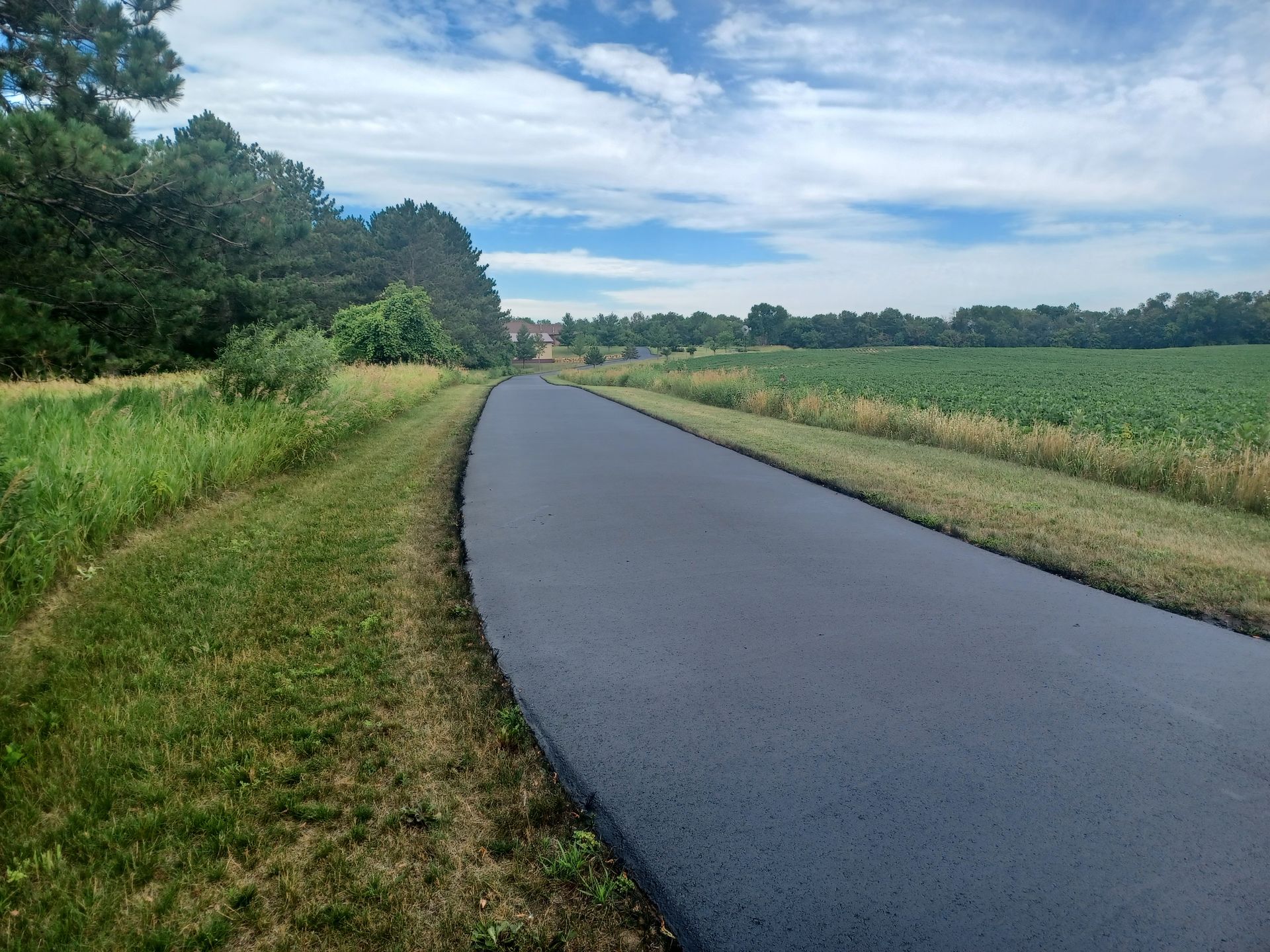 Curving paved road beside green fields and trees under a cloudy blue sky