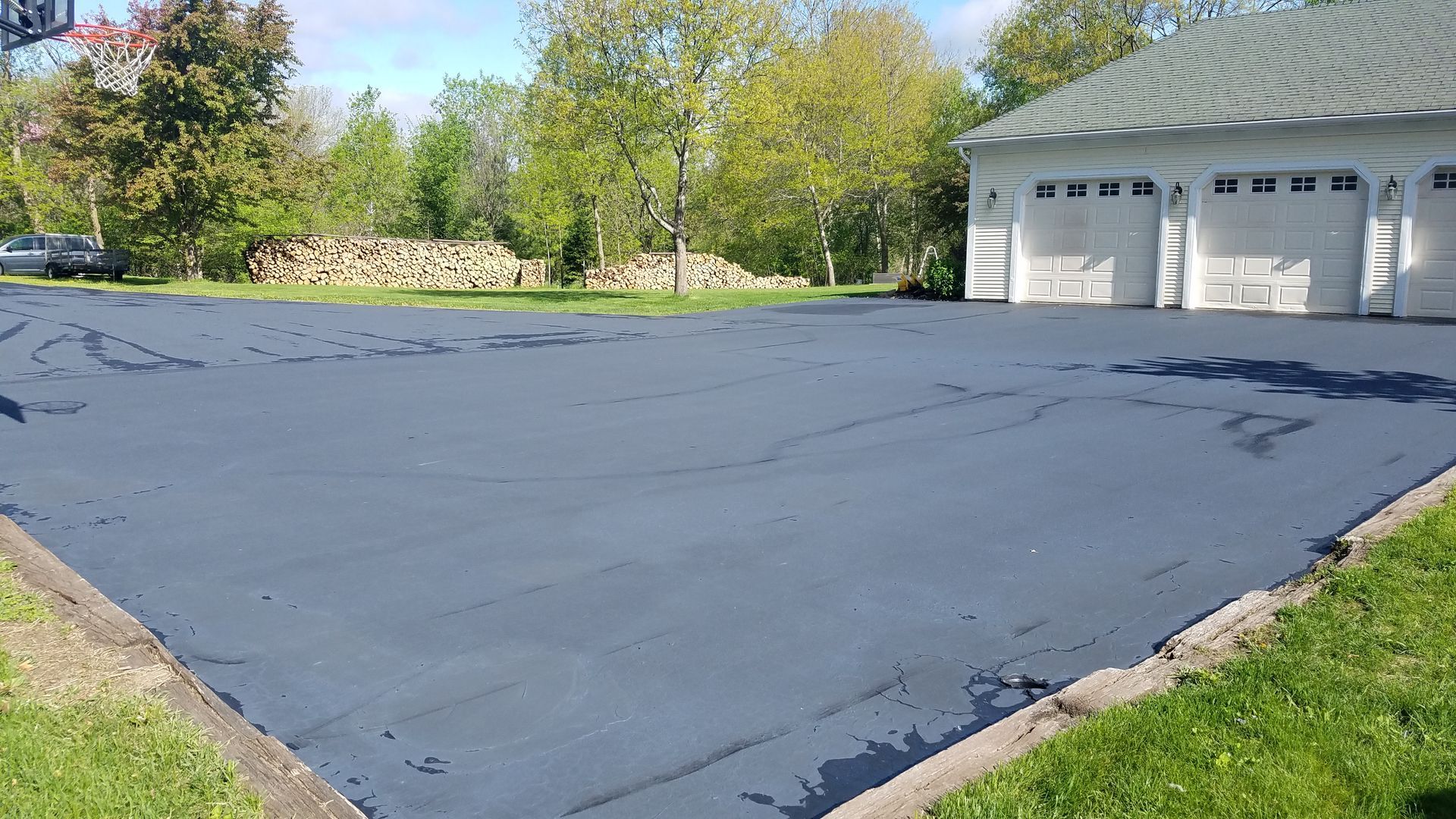 Freshly paved black driveway beside a white garage, with trees and grass in the background.