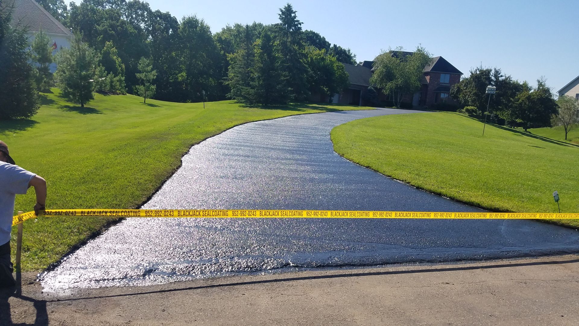 Curving paved path through green grass and trees on a sunny day, with a yellow barrier across the foreground