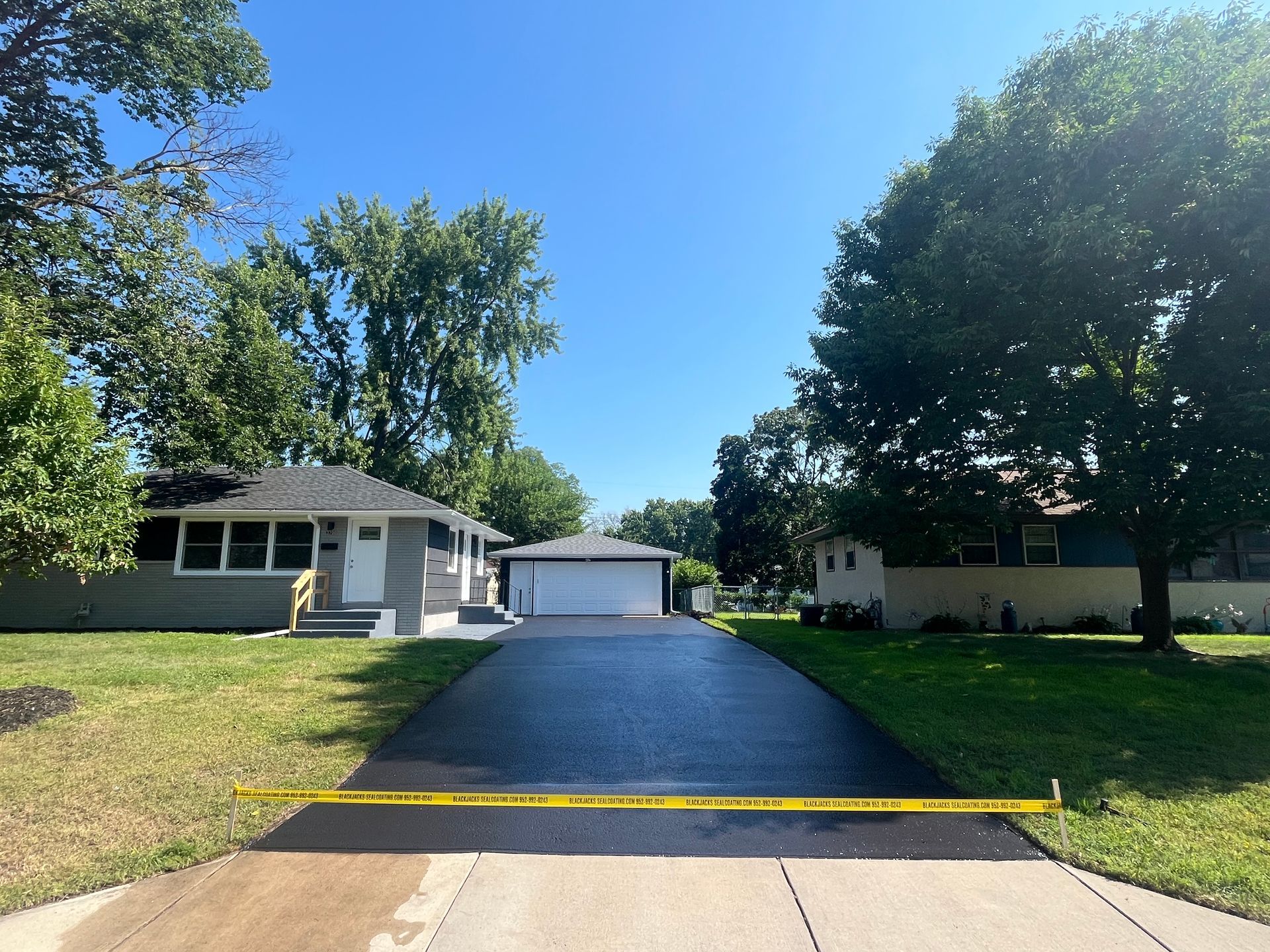 Residential driveway with fresh black pavement leading to a garage between trees and grassy lawns