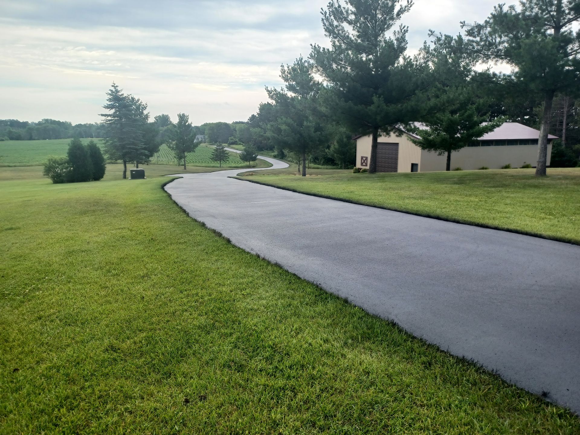 Curving wet driveway through a green lawn near trees and a house under cloudy skies