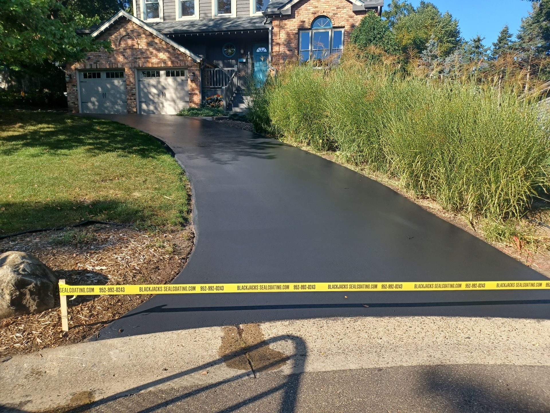 A newly paved driveway leads to a house, blocked by yellow caution tape and bordered by tall grass.