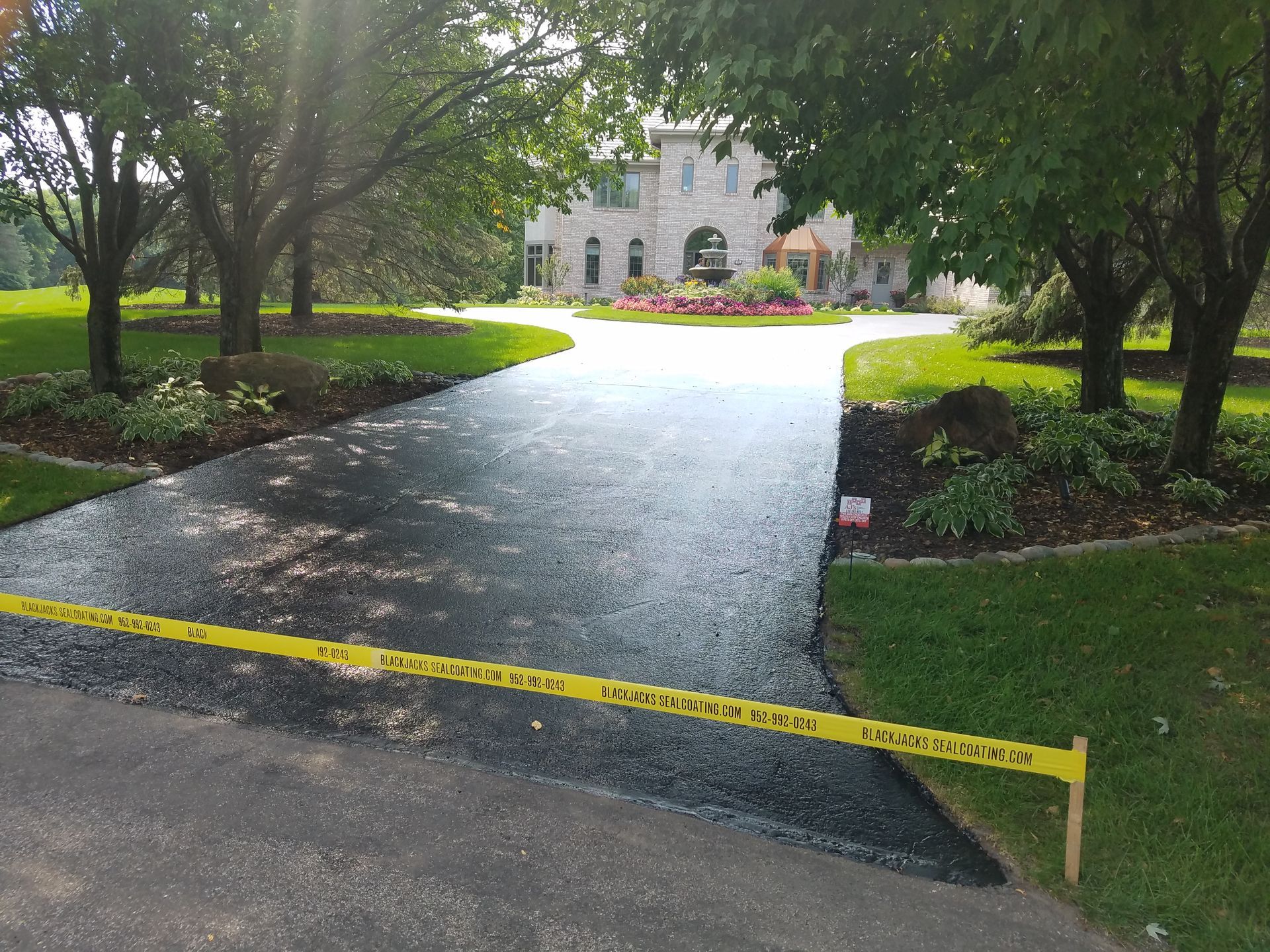Caution tape blocks a wet driveway leading to a stone building surrounded by trees and landscaping.