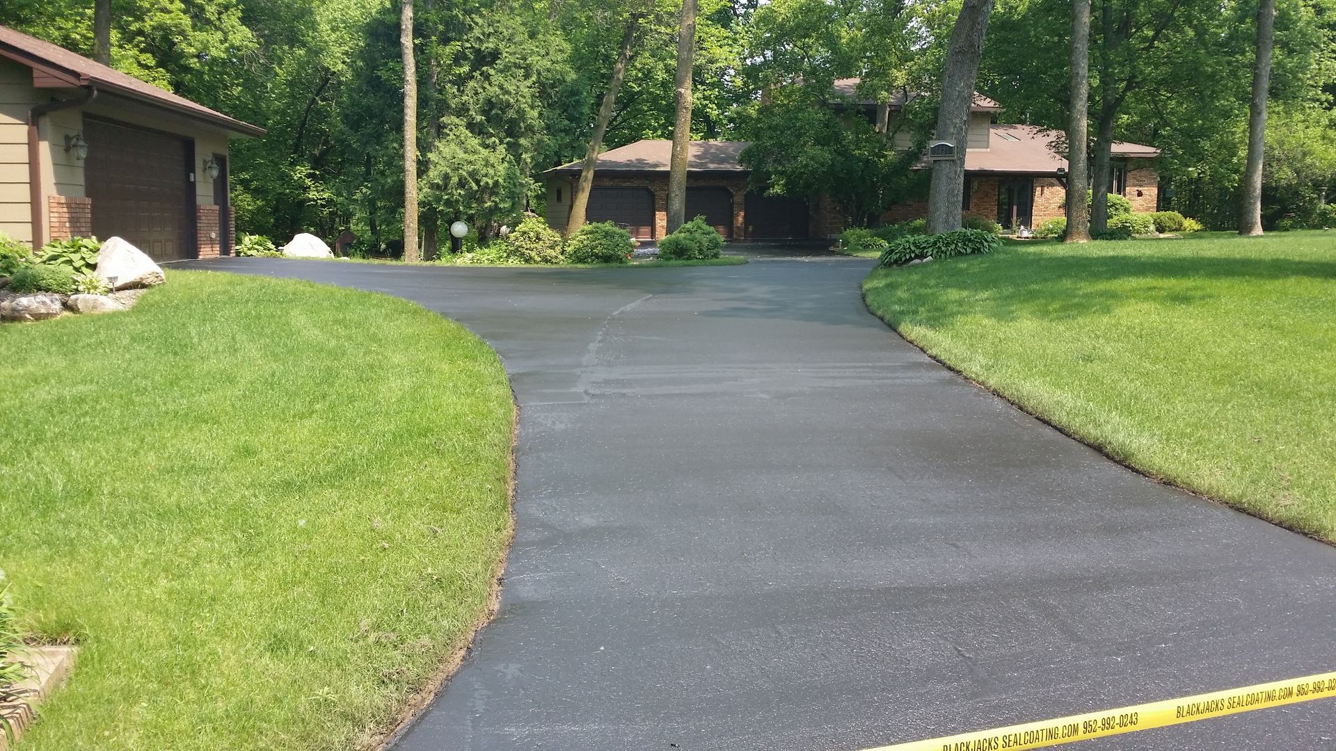 Curved paved driveway in a wooded residential area, with brick houses and green lawns on both sides.