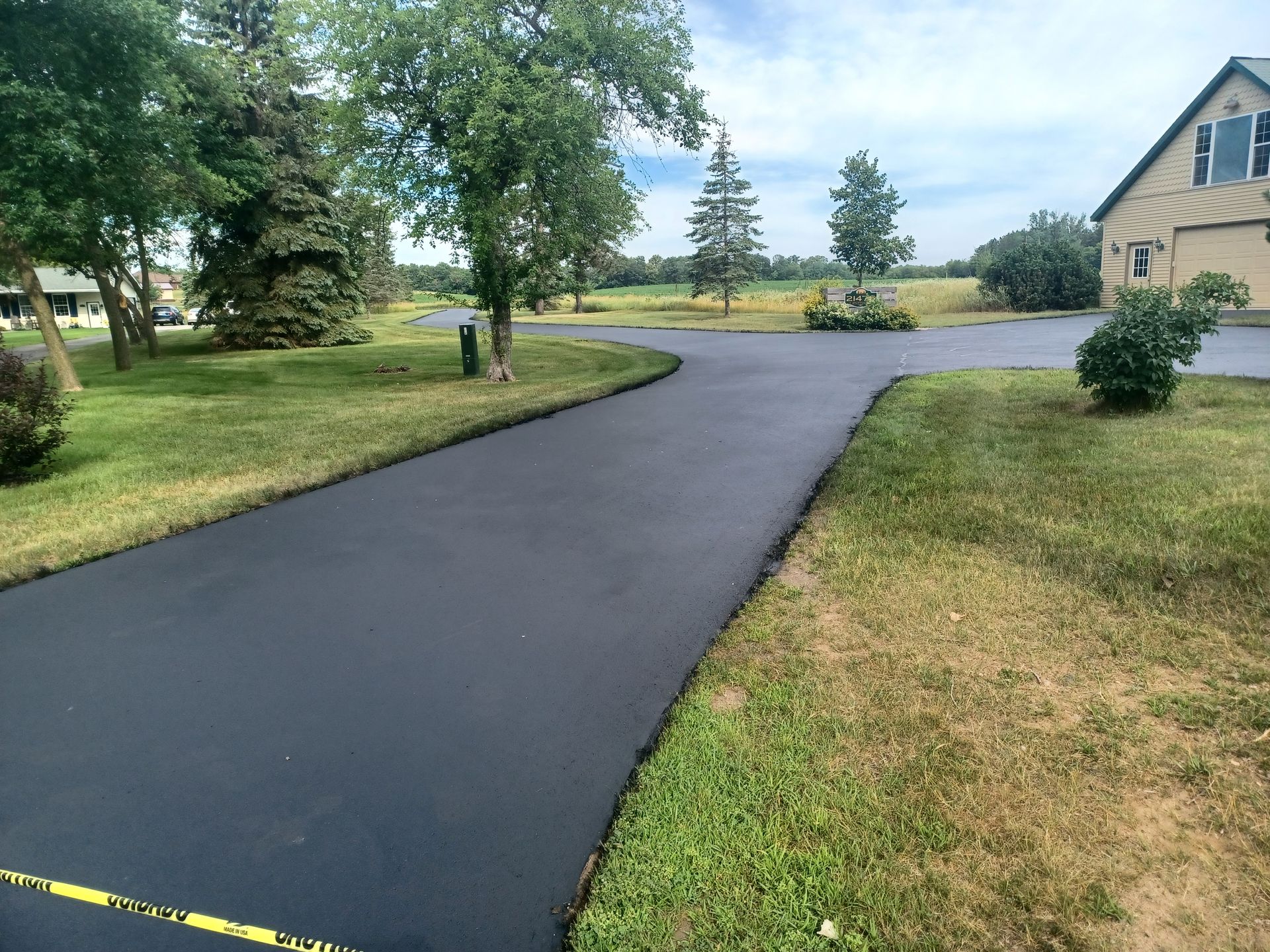 Fresh black asphalt driveway curving through a grassy yard beside a house and trees