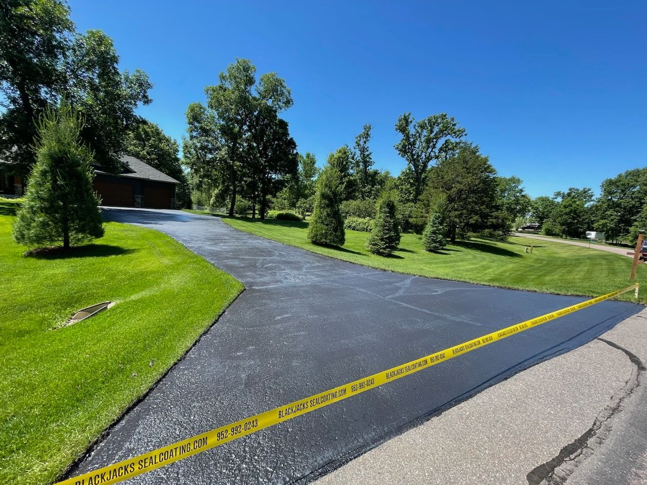 New blacktop driveway at a grassy property, bordered by yellow caution tape and trees under a blue sky
