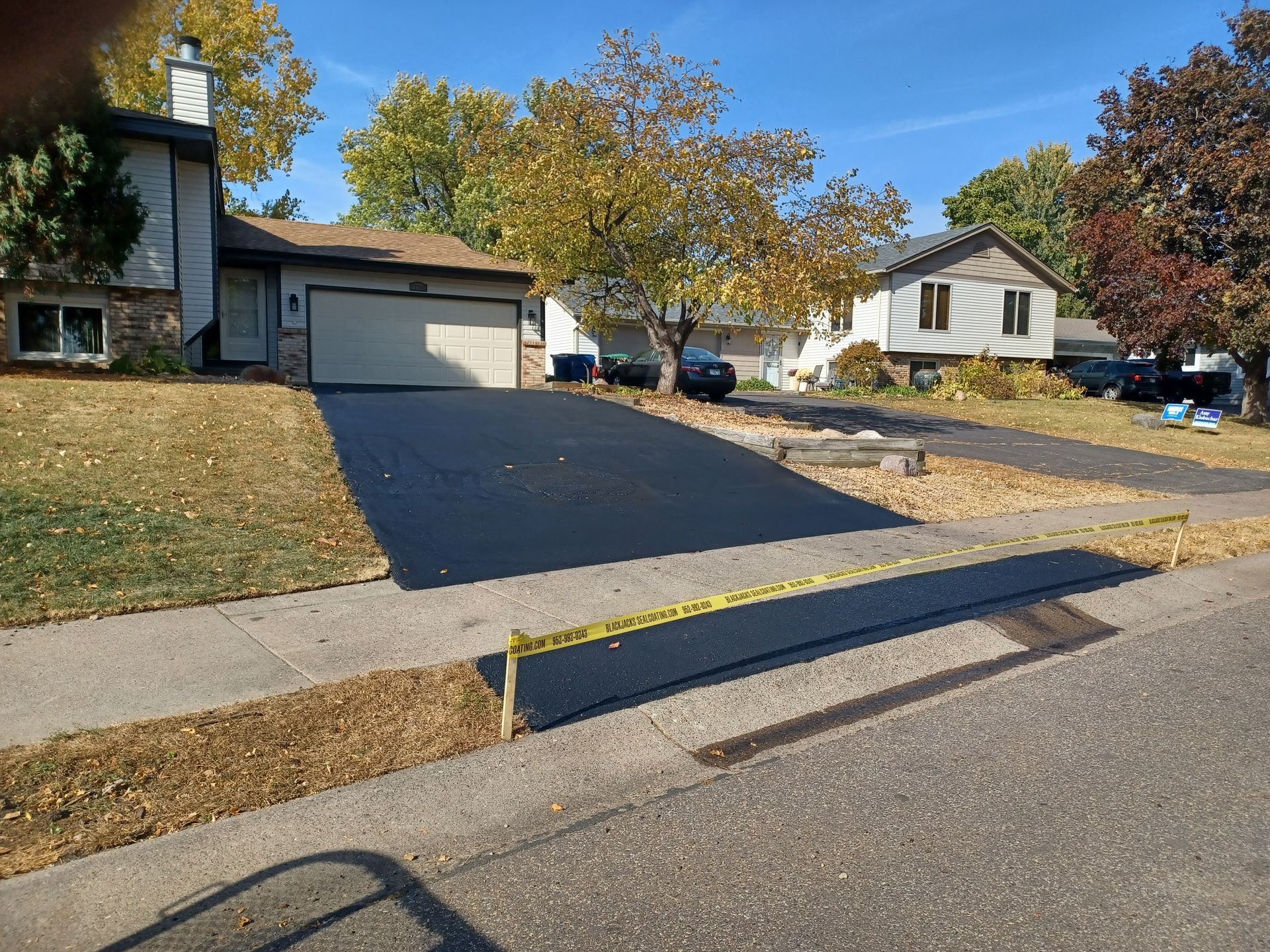 Suburban driveway being repaved with fresh black asphalt beside a curb and houses in fall.