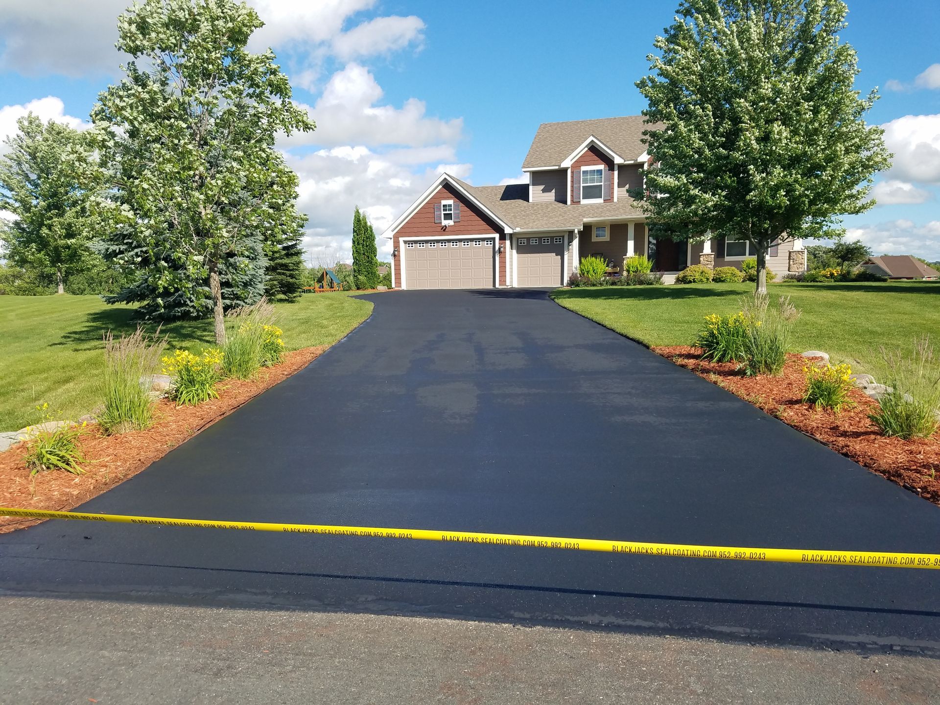 Freshly paved black driveway leading to a suburban house with garage and trees under a blue sky