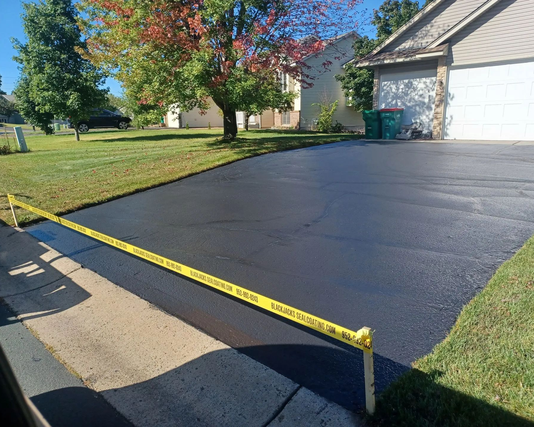 Freshly paved black driveway beside a garage, blocked by yellow caution tape in a suburban yard.