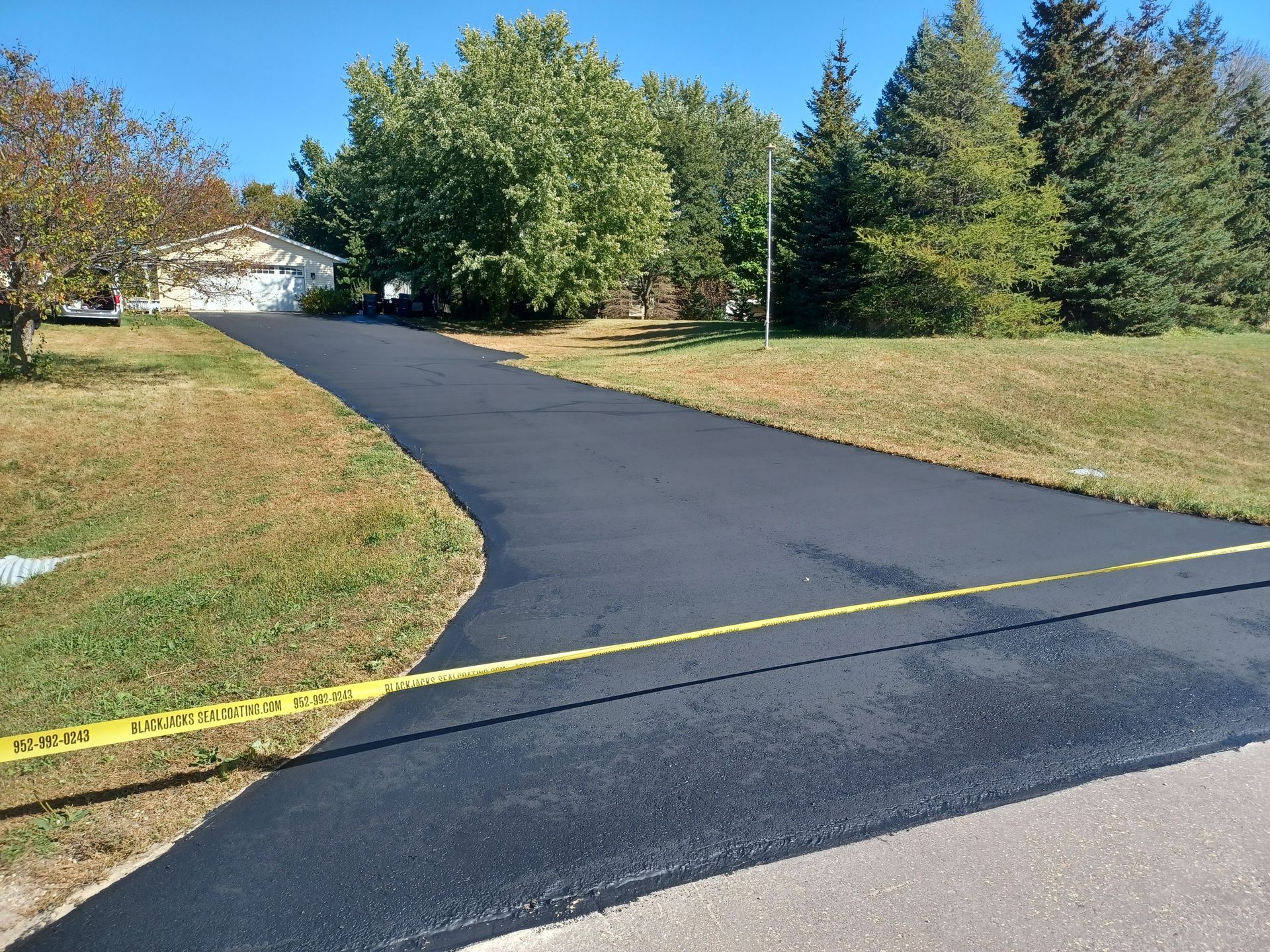 Fresh black asphalt driveway curving through a grassy yard, bordered by yellow caution tape and trees