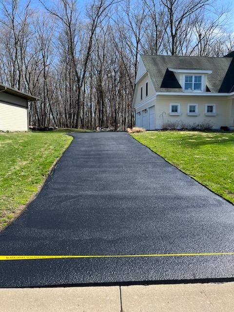 Fresh black asphalt driveway leading to a white house, bordered by green grass and leafless trees