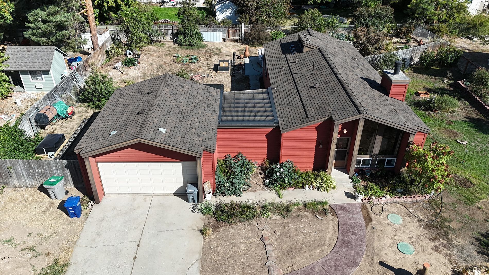 Red house with garage and a dark roof. Concrete driveway, front yard, and plants.