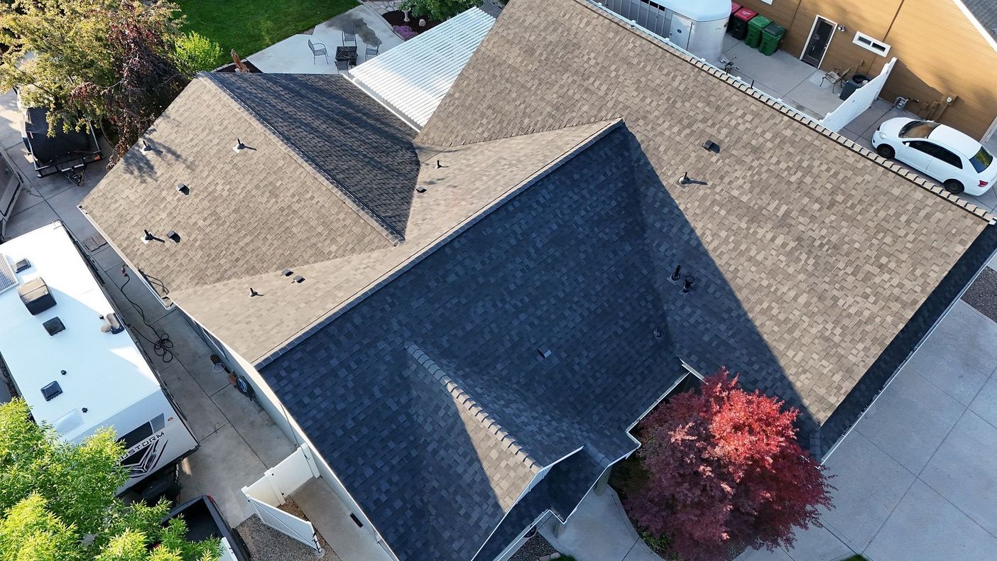 Aerial view of a suburban house with a gray roof, driveway, parked vehicles, and a red tree in the yard