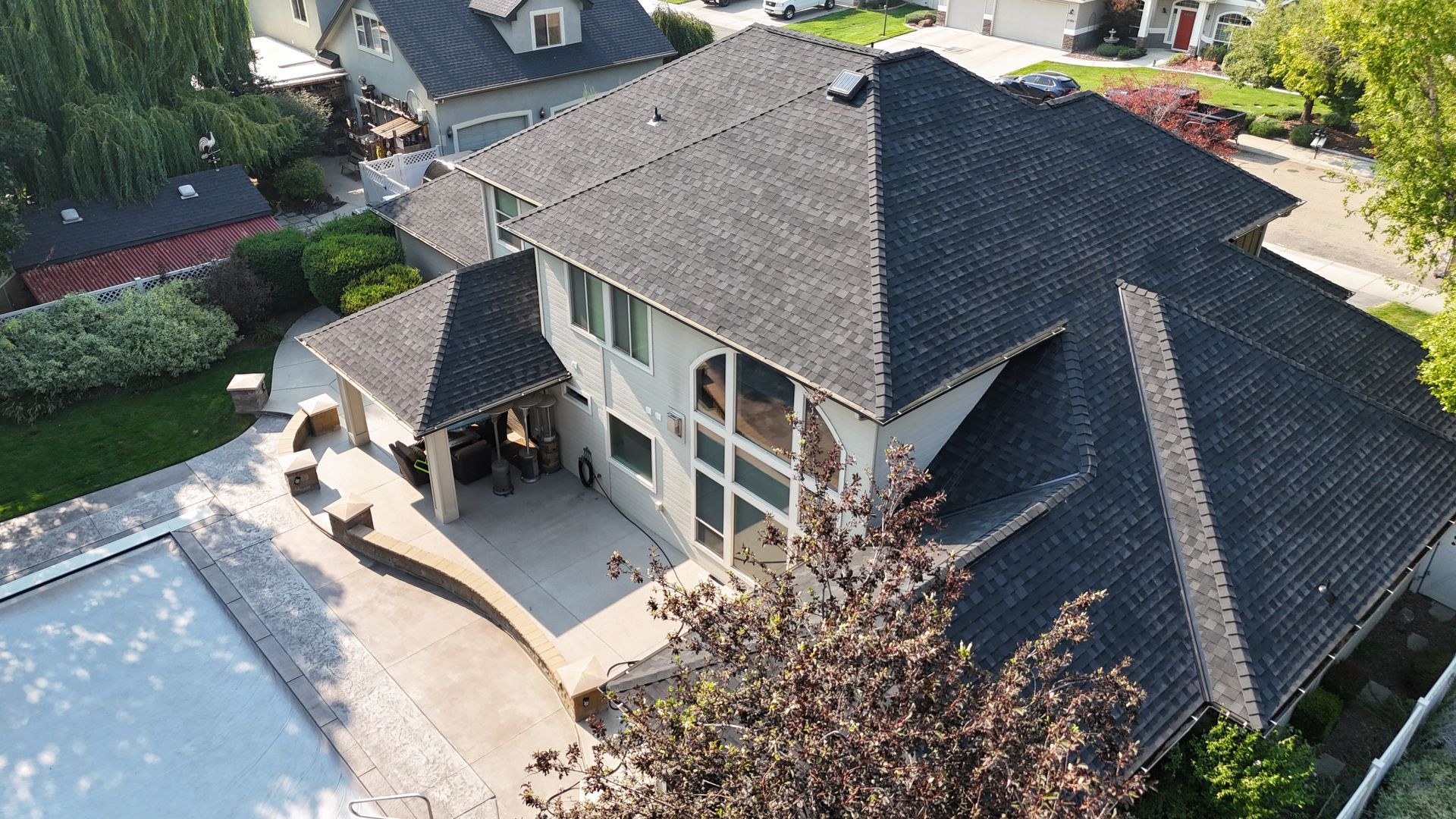 Aerial view of a house with a dark shingled roof, patio, and backyard in a suburban neighborhood