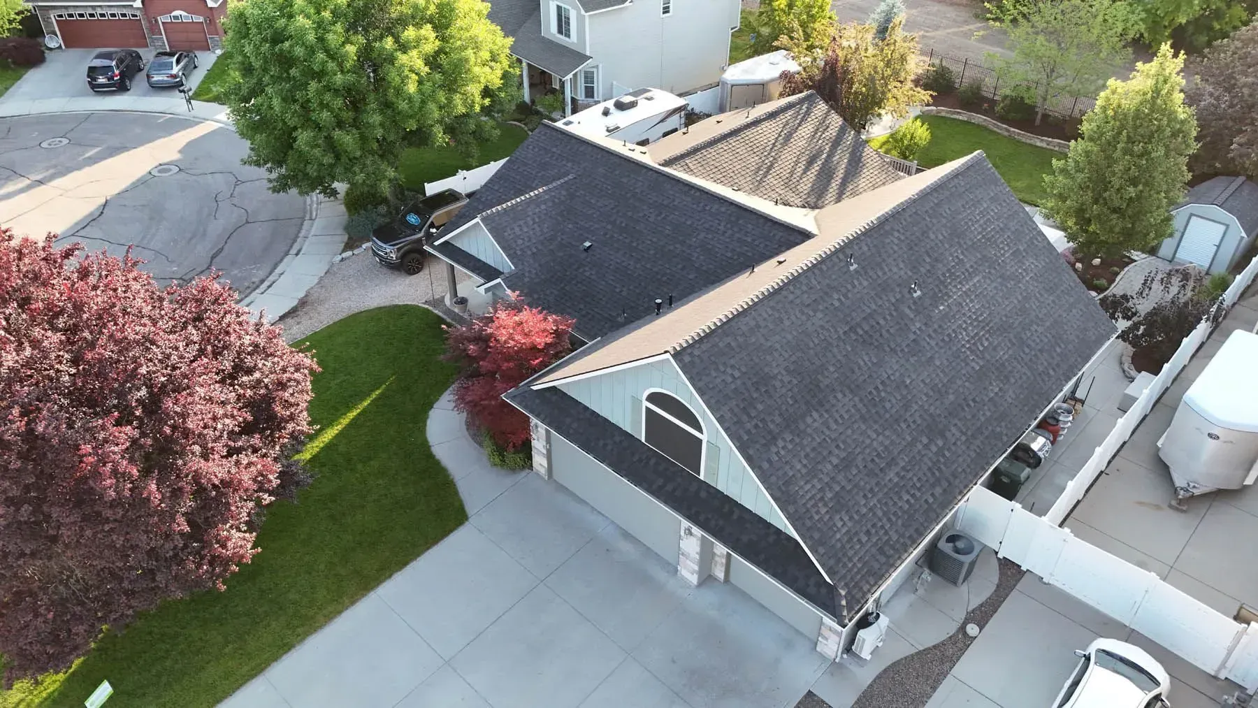 Aerial view of a gray house with a dark roof and a paved driveway, surrounded by green grass and trees.
