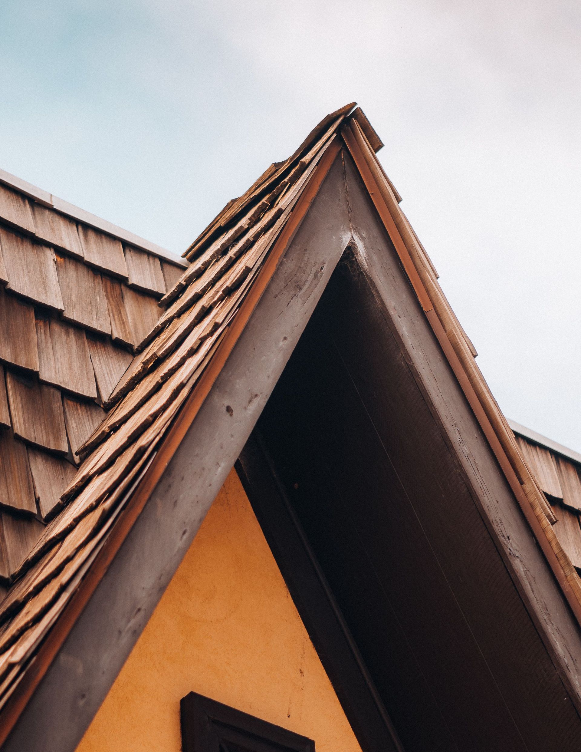 Close-up of a peaked roof with brown shingles and an orange wall against a cloudy sky