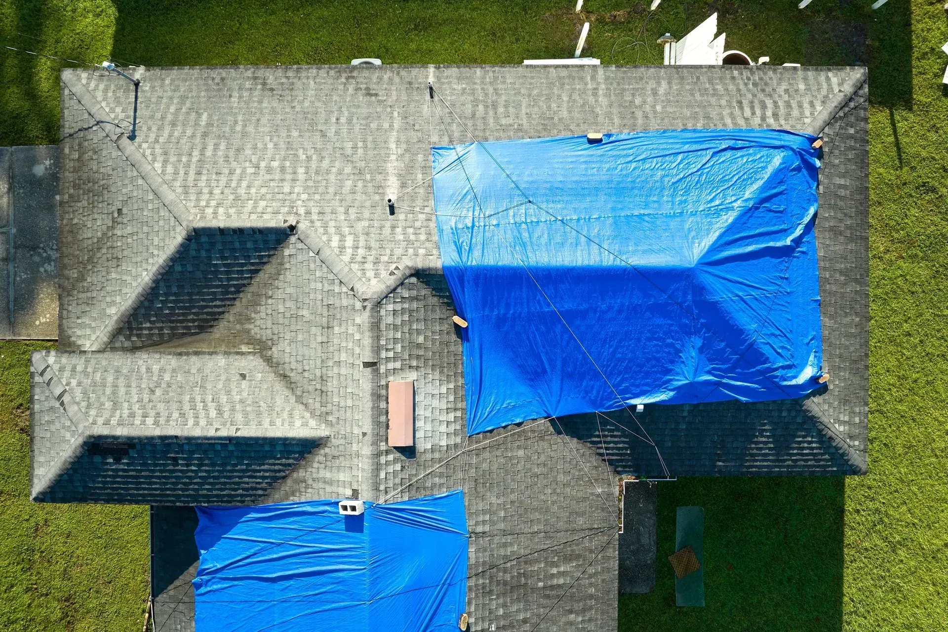 Overhead view of a house roof with blue tarps covering damaged sections; gray shingles, green lawn.