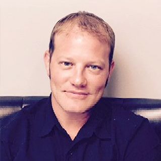 A man in a black shirt is sitting on a couch and smiling for the camera.