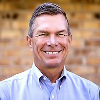 A man in a blue shirt is smiling in front of a brick wall.