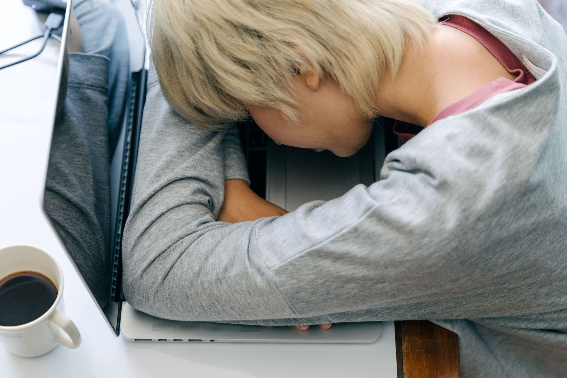 Person sleeping on a desk beside a laptop and a cup of coffee