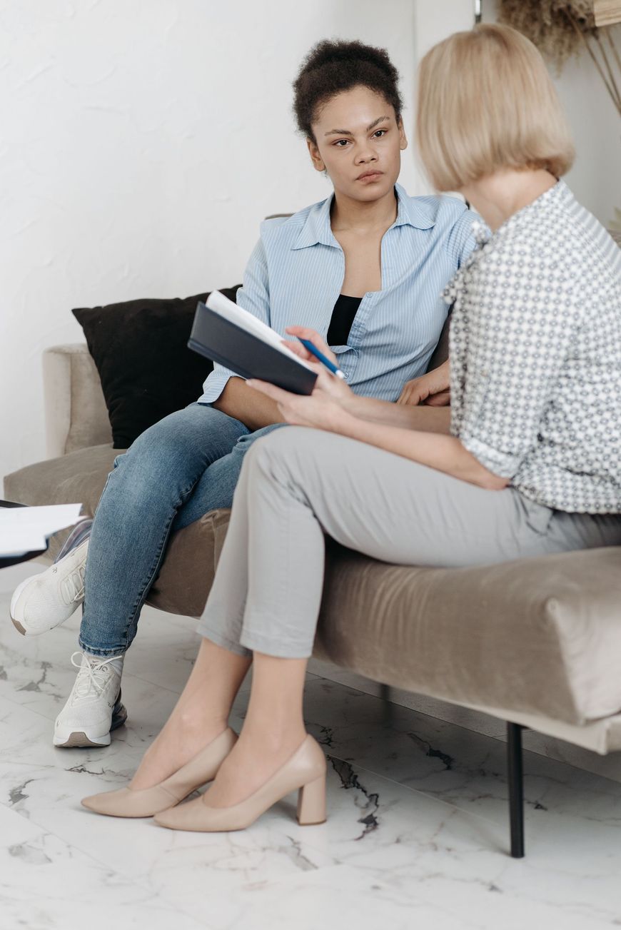 Two women talking on a couch, one holding a notebook in a bright living room.