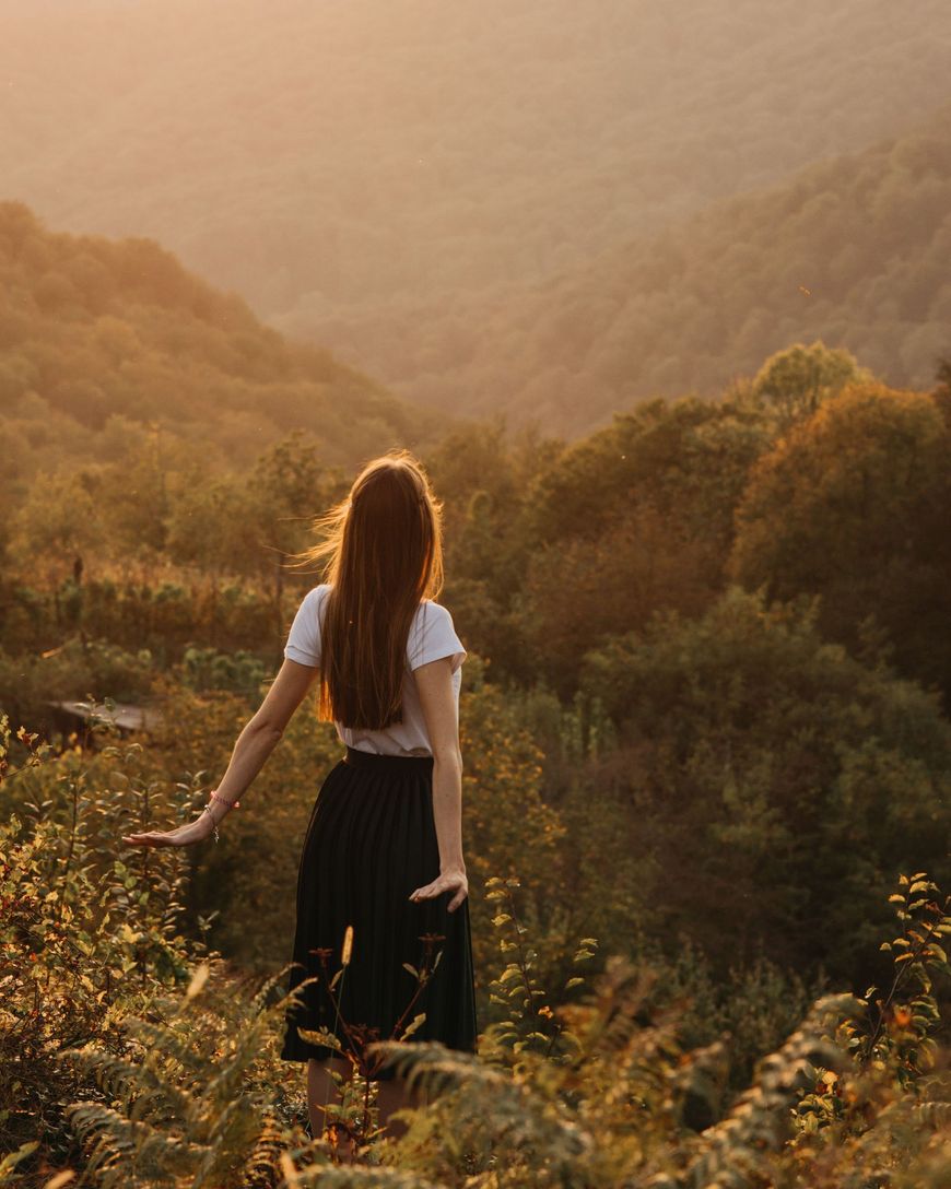 Person with long hair standing in a sunlit hillside meadow overlooking a forested valley