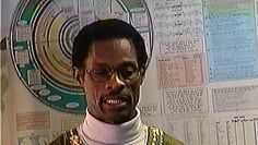 A man wearing glasses and a turtleneck is standing in front of a wall.