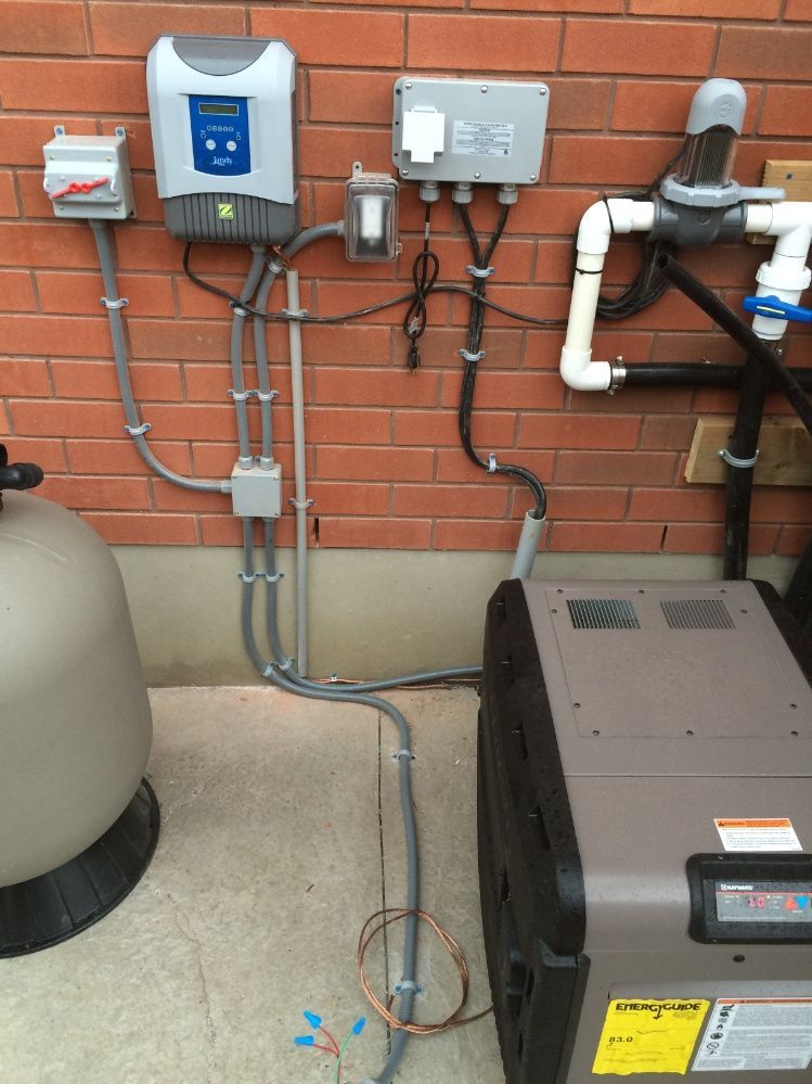 Pool pump and heater equipment mounted on a brick wall, with various electrical conduits and control boxes attached.