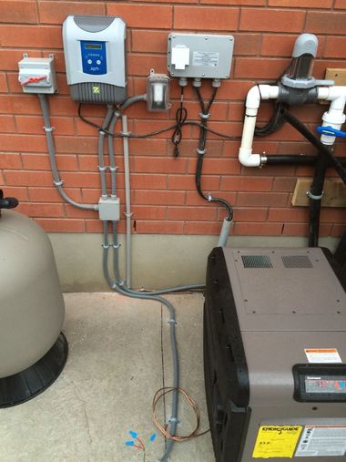Pool pump and heater equipment mounted on a brick wall, with various electrical conduits and control boxes attached.