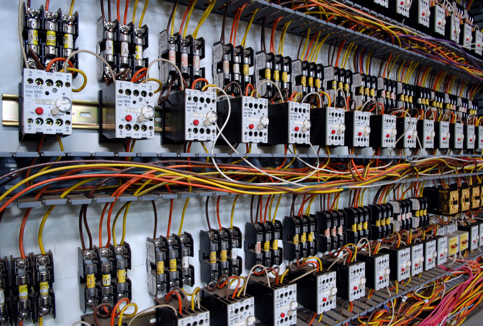 An electrical control panel with rows of circuit breakers, relays, and organized wiring on a light-colored backboard.