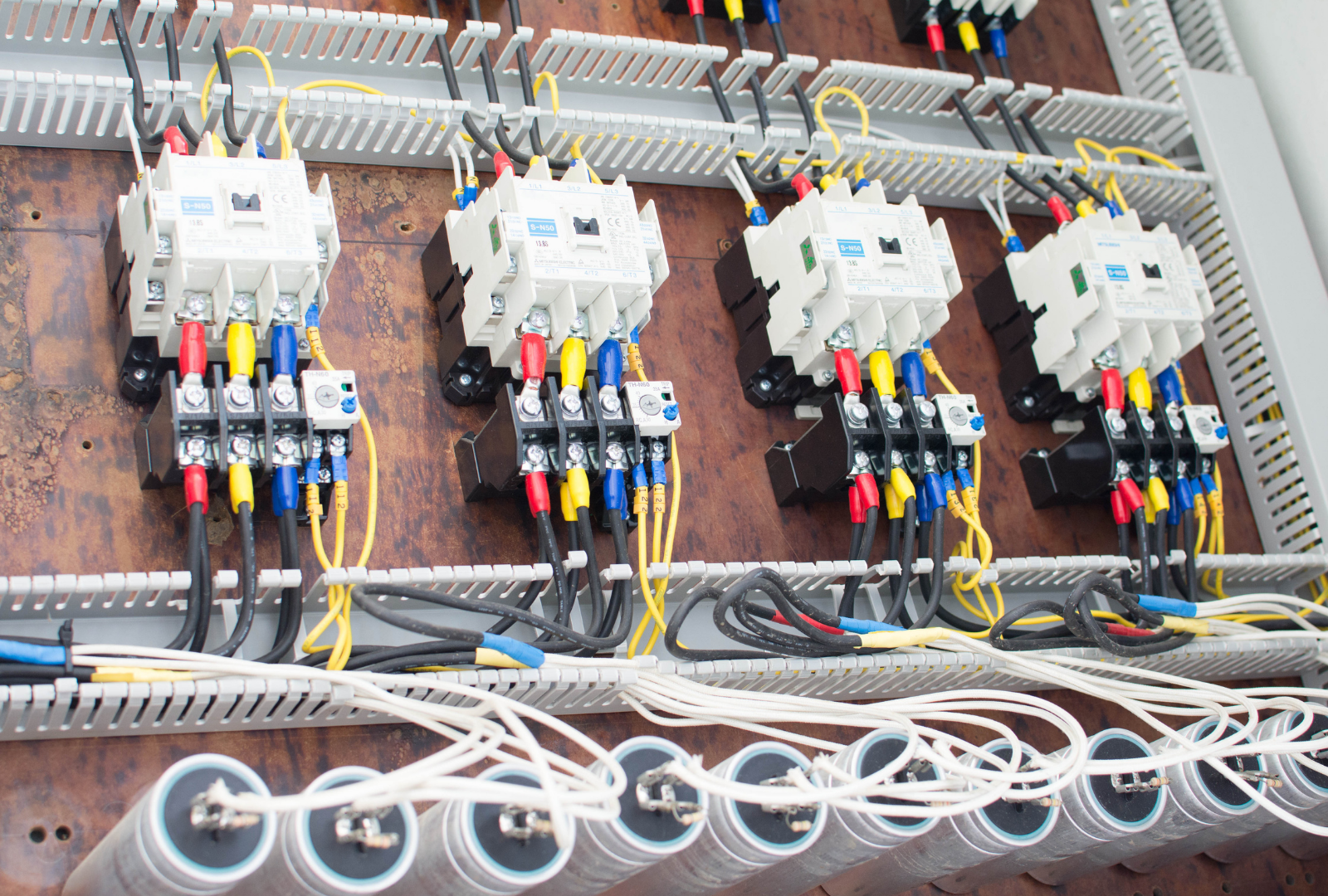 Electrical control panel with four contactors wired to capacitors, mounted on a wooden backboard inside a cabinet.