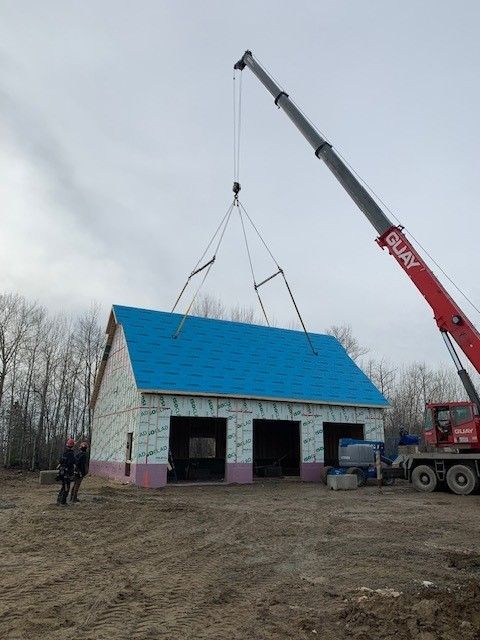 Une grande grue rouge soulève une structure préfabriquée au toit bleu et la dépose sur un chantier où se tiennent deux ouvriers à proximité.
