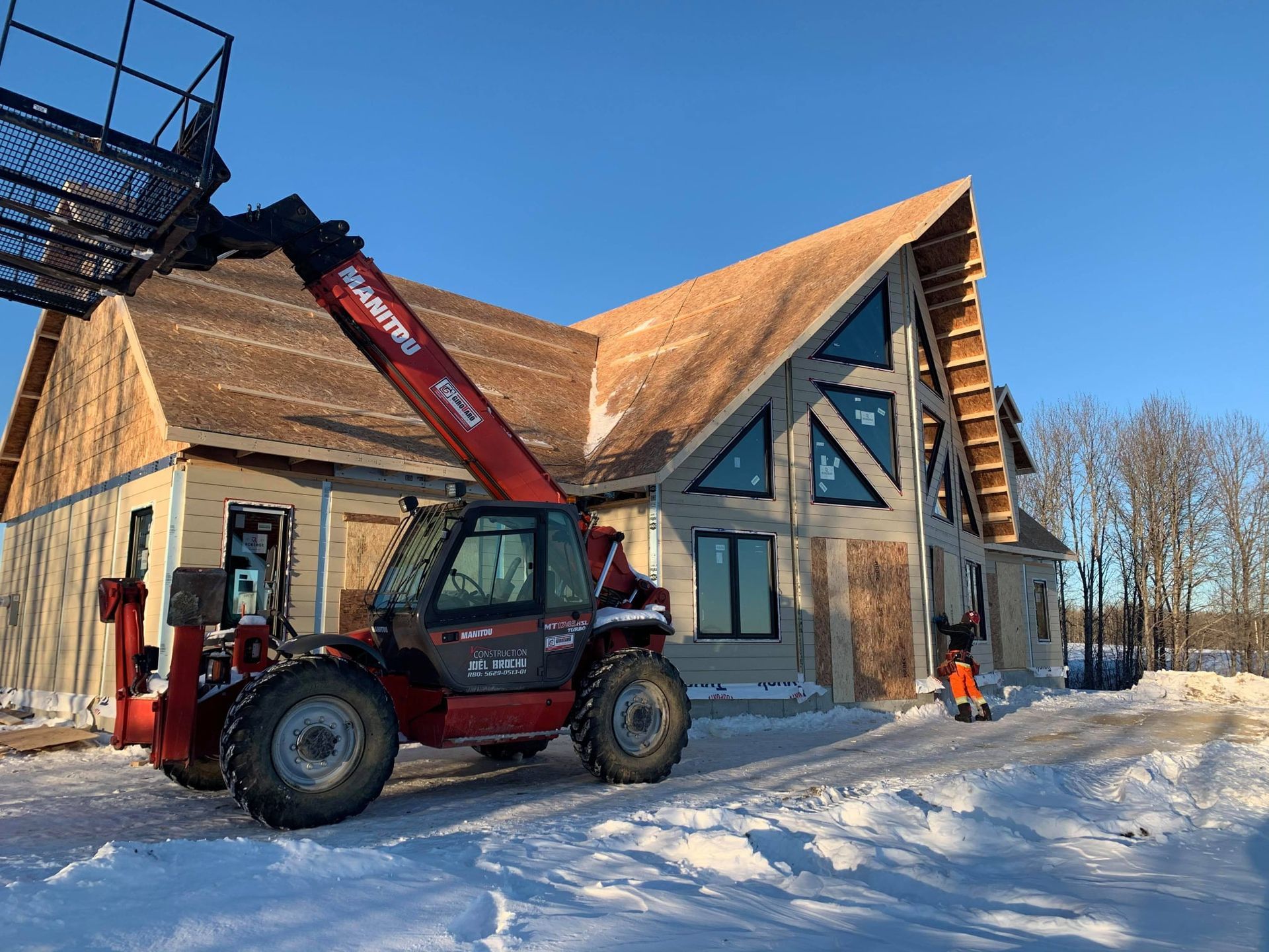 Un chariot télescopique Manitou rouge est stationné dans la neige devant une maison en construction avec une grande fenêtre triangulaire.
