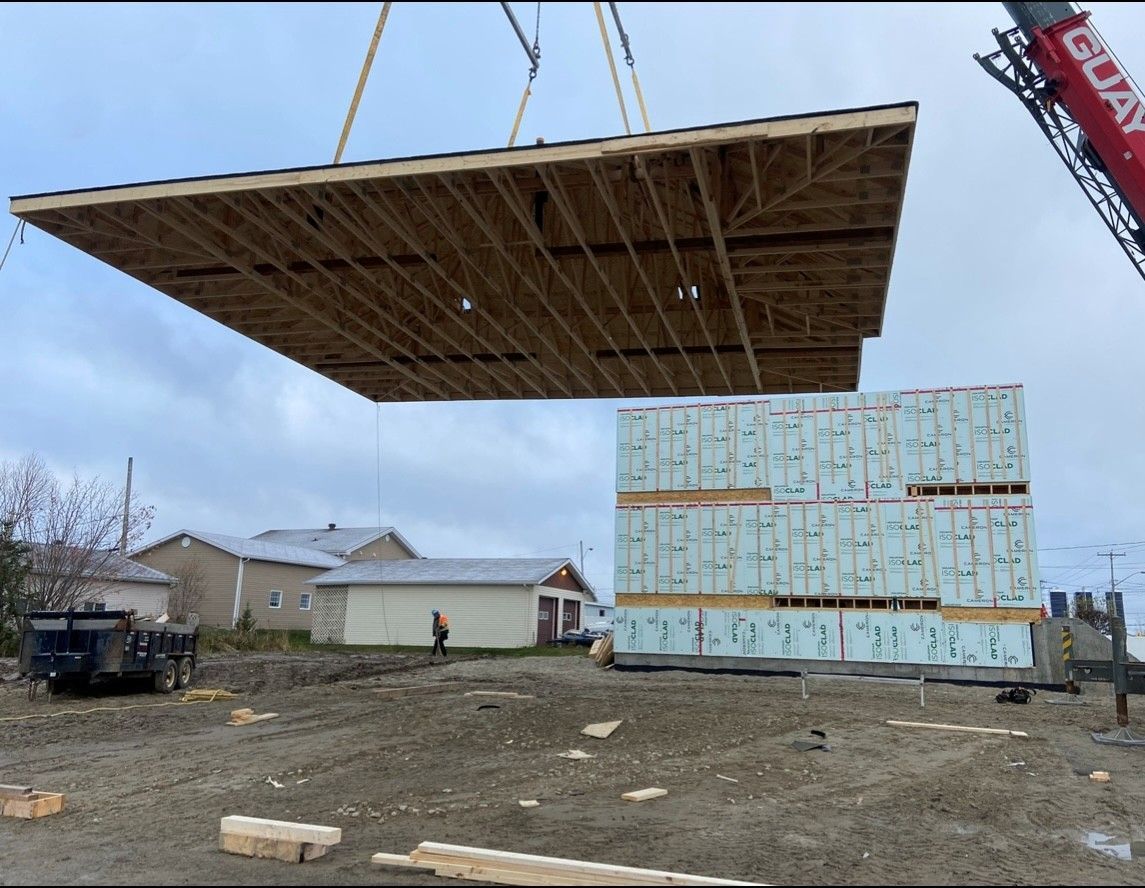 Une grue dépose une grande structure à toit plat sur une maison partiellement construite, recouverte de panneaux isolants bleus, sur un terrain extérieur.