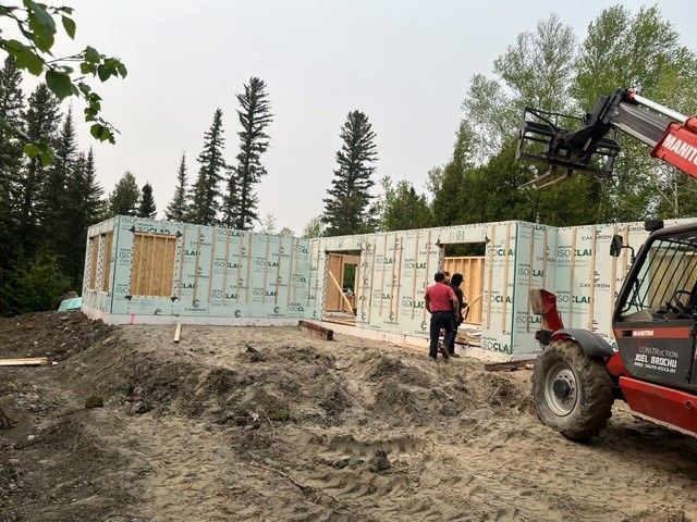 Deux ouvriers se tiennent près des murs isolés bleu clair, partiellement construits, d'une maison dont la structure est en place dans une zone boisée, à l'aide d'un chariot élévateur.