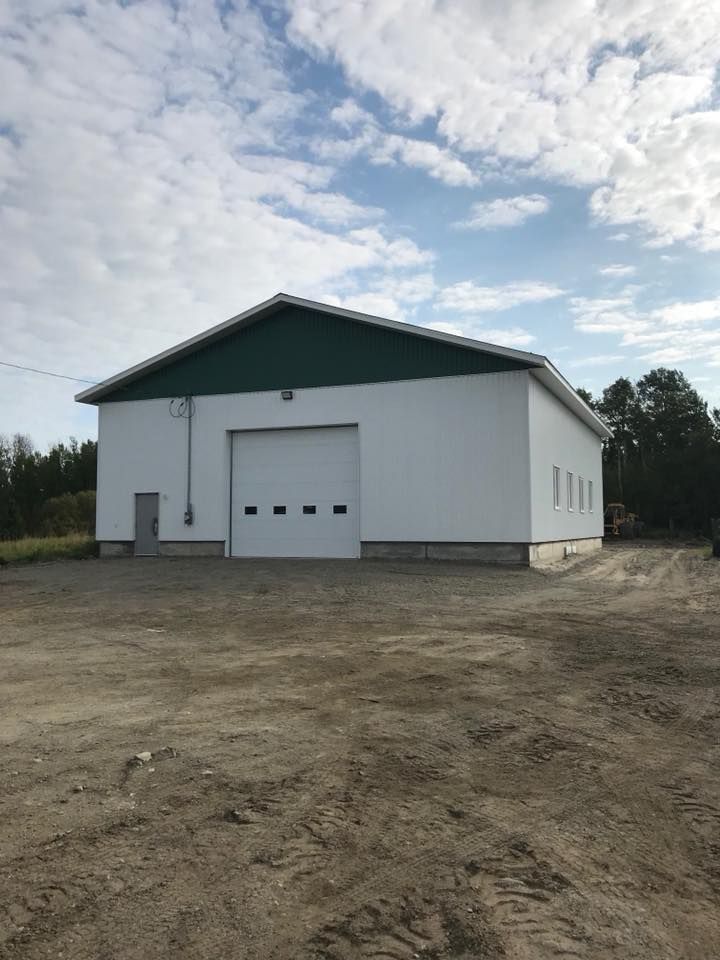 Un garage blanc indépendant, avec un toit à pignon vert et une porte sectionnelle centrale, sur un terrain en terre battue sous un ciel nuageux.