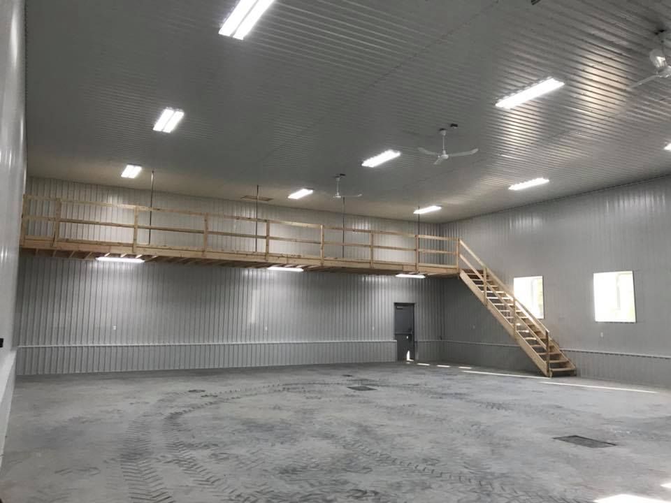 Un grand espace de travail industriel vide, avec des murs métalliques, un sol en béton, un éclairage zénithal et une mezzanine en bois avec escalier.