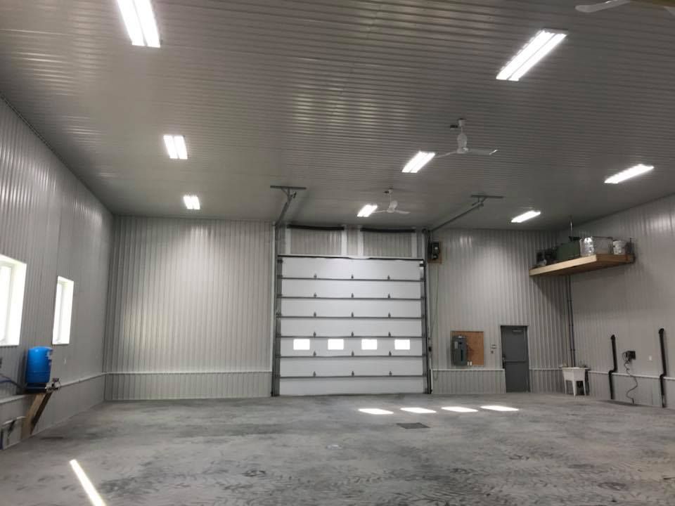 Un grand garage industriel vide, avec des murs en panneaux métalliques, une grande porte sectionnelle, des éclairages au plafond et un sol en béton.
