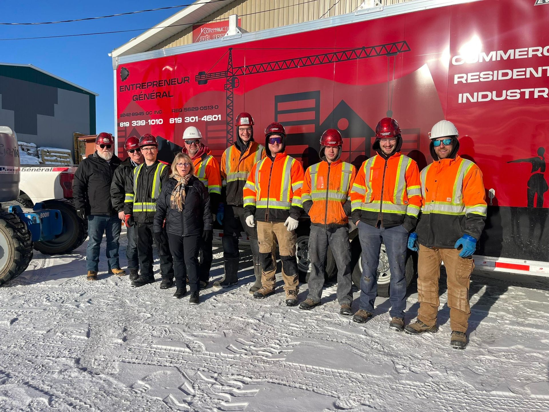 Un groupe d'ouvriers du bâtiment, vêtus de gilets orange haute visibilité et de casques de chantier, se tiennent devant une remorque utilitaire rouge.